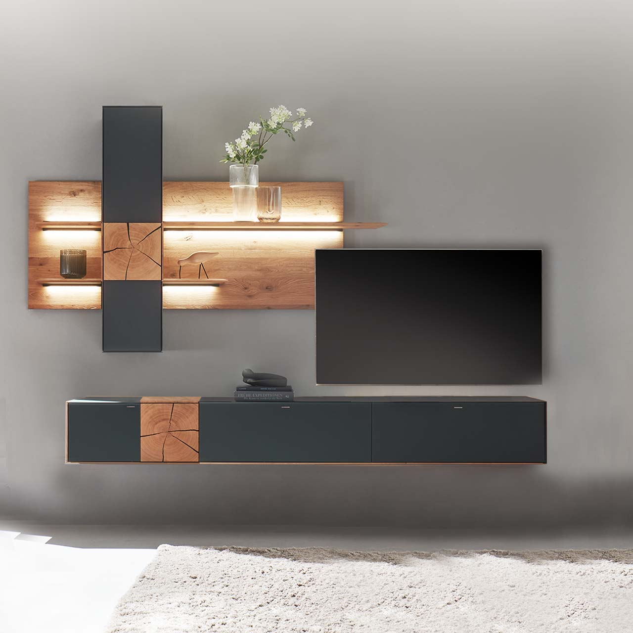 Moderne Wohnwand aus massiver Kerneiche mit anthrazitfarbenen Fronten. Unten ein langes Hängelowboard mit dekorativer Holzapplikation, oben eine Wandpaneel-Kombination mit Regalböden, Schrank und indirekter Beleuchtung, dazwischen hängender Fernseher.