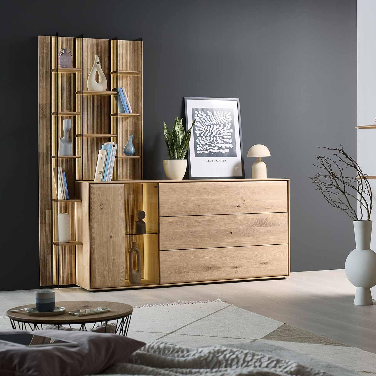 Modernes Sideboard aus massiver Kerneiche Natur mit beleuchteter Glas/Holztür, einer Holzklappe oben und zwei Holzschubkästen. Das Sideboard ist integriert in ein links stehendes, hohes Wandregal mit Holzböden und Glasseiten. - im Wohnzimmer stehend.