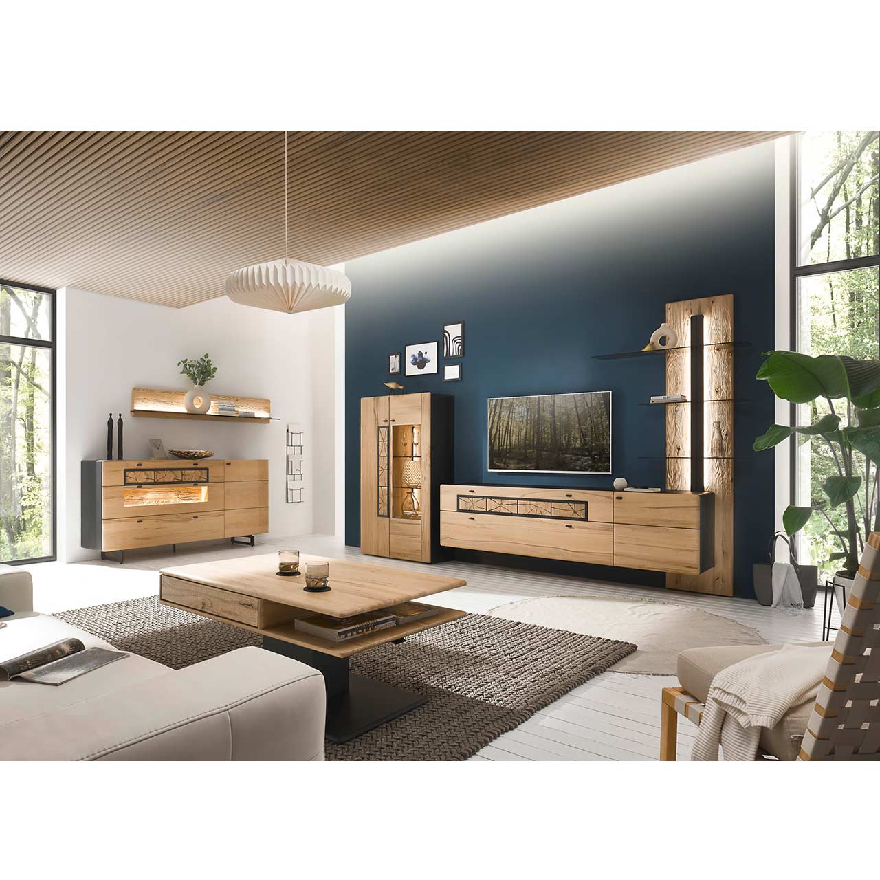 Eingerichtetes Wohnzimmer mit moderner Wohnwand aus Kernbuche vor einer dunkelblauen Wand bestehend aus Highboard, Hängelowboard und Paneel. Links das passende Sideboard auf Metallkufen darüber ein Regal. Im Vordergrund der passende Couchtisch.