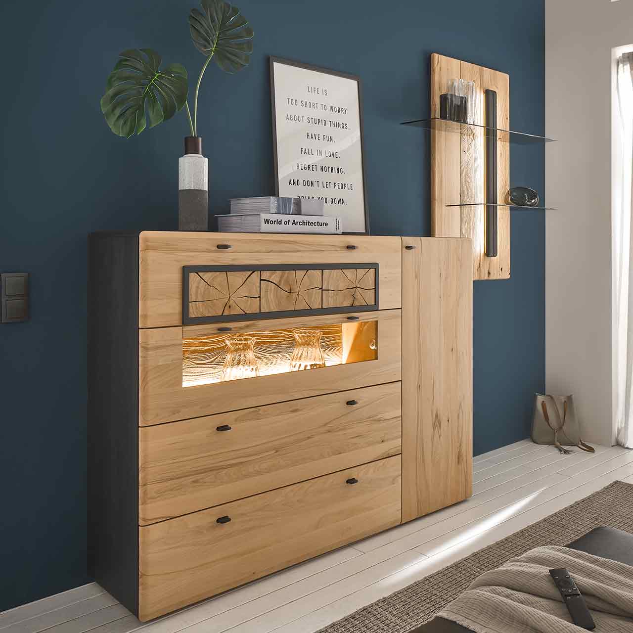 Vor dunkelblauer Wand stehendes Highboard aus heller Kernbuche. Mit Holztür und drei Schubkästen, oben verziert mit dekorativen Hirnholzscheiben dazwischen eine Klappe mit Glasausschnitt. Korpus seitlich Anthrazit lackiert. Daneben ein Wandpaneel.