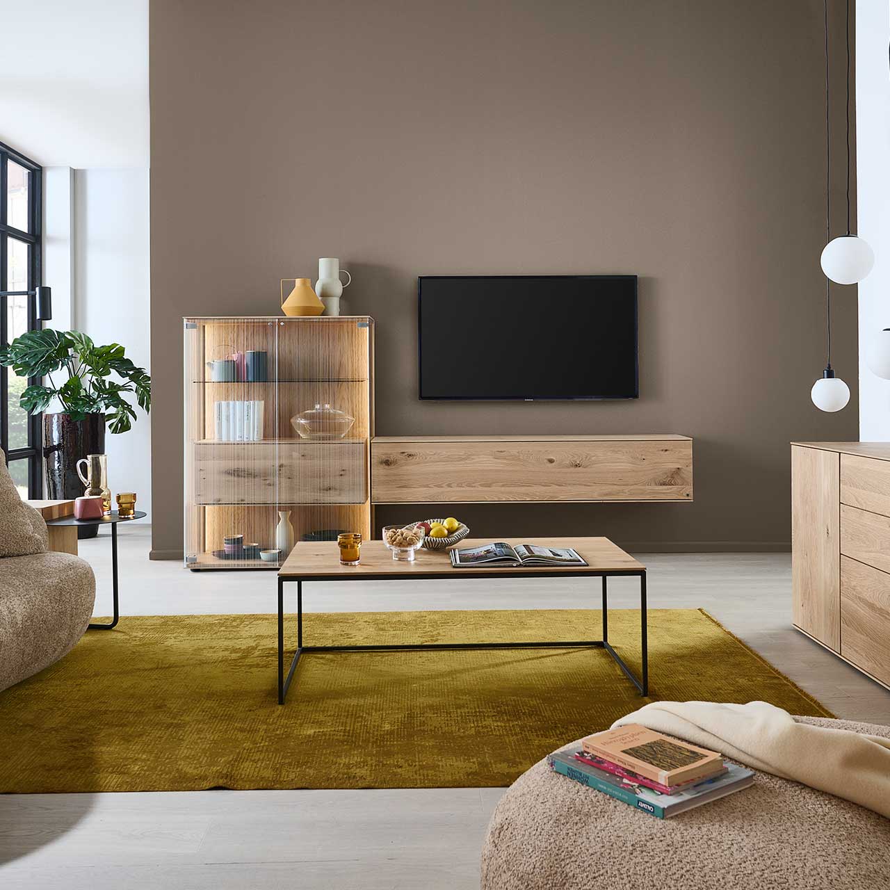 Wohnzimmer mit Fernseher und zweiteiliger Wohnwand aus Kerneiche Natur: Highboard mit satinierten Glastüren und innenliegendem Holzschrank und schlichtes Hängelowboard. Davor passender rechteckiger Couchtisch mit Metallgestell.