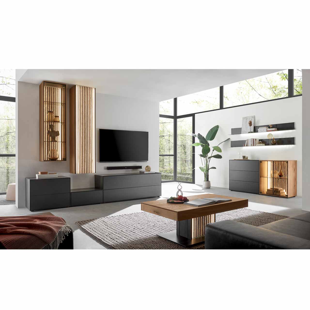 Modern eingerichtetes Wohnzimmer mit fünfteiliger Massivholz-Wohnwand bestehend aus Lowborads in anthrazit, Hängevitrine und Hängeschrank in hellem Holz mit Lamellen. Davor der passende helle Couchtisch. Rechts ein Sideboard und Wandregale.
