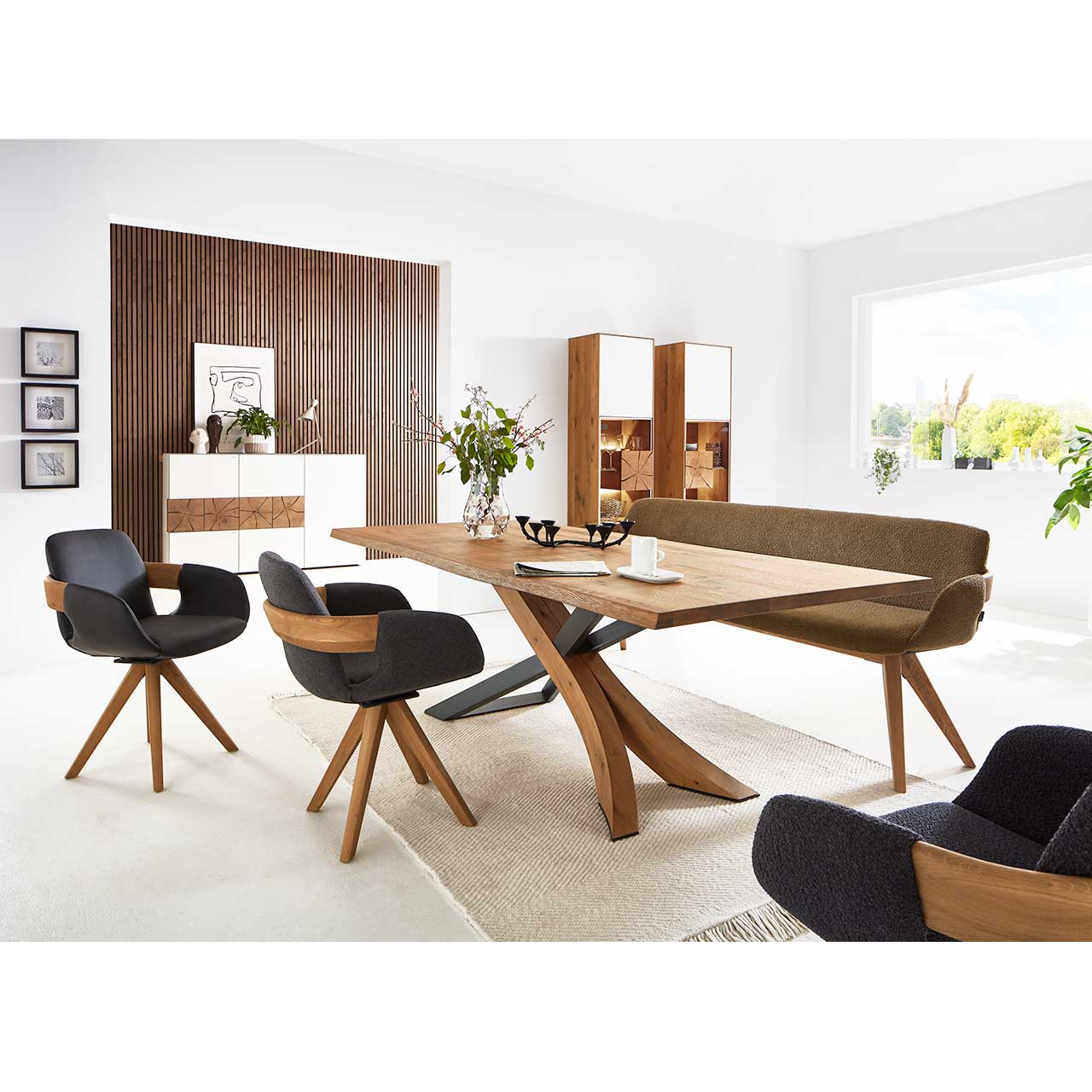Modernes Esszimmer mit massivem, rechteckigen Holztisch, umgeben von zwei Stühlen mit dunklem Bezug auf Holzgestell und einer Sitzbank. Hinten an der Wand stehend die passenden Esszimmermöbel.