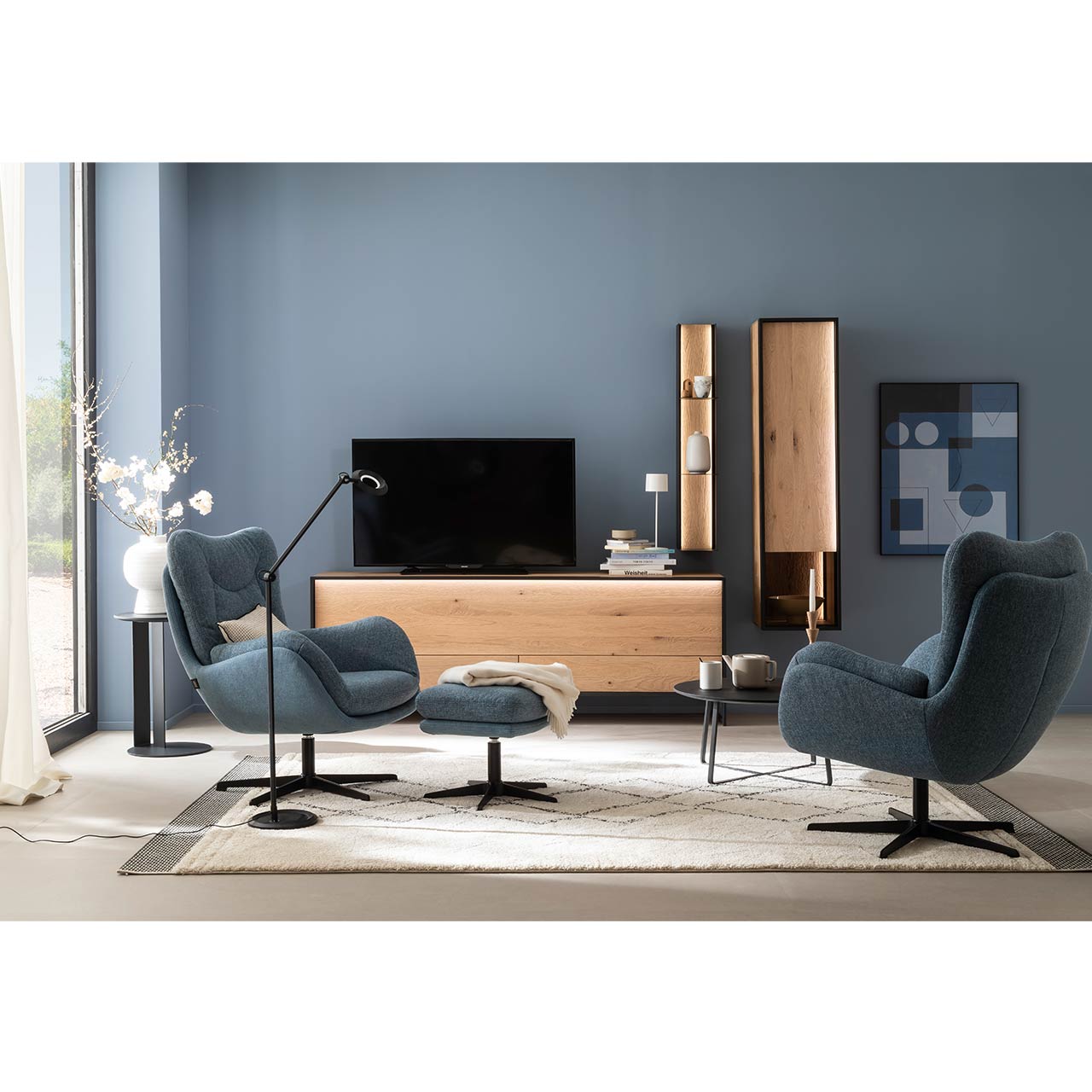 Eingerichtetes Wohnzimmer mit blauen Wänden und einer dreiteiligen Wohnwand aus massiver Kerneiche Natur bestehend aus einem schlichten Lowboard auf Metallkufen, einem Hängeschrank mit Glasausschnitt und einem Metallregal mit Riffholz.